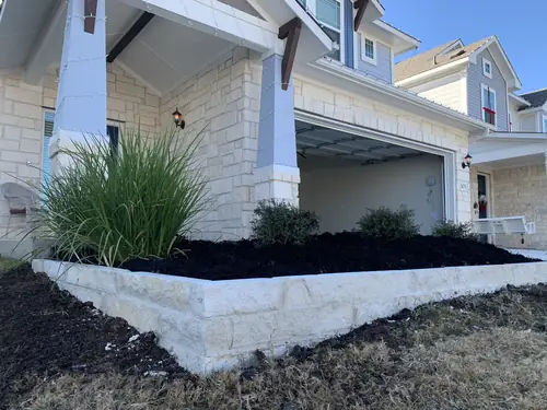 Limestone-block raised planting bed with fresh mulch and ornamental grasses across a stone-front home's entry and garage
